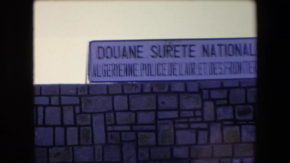 1971 Algeria 8mm frame showing a Douane Surete Nationale Algerienne Police de l'Air et des Frontieres sign mounted above a patterned stone wall.