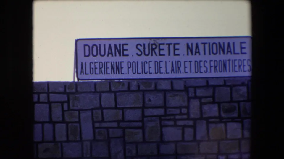 1971 Algeria 8mm frame with the French-language Douane Surete Nationale Algerienne sign visible over a stone wall, cropped by the film frame edge.