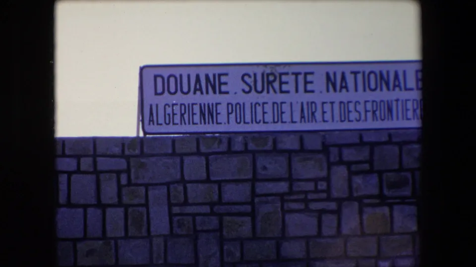 1971 Algeria 8mm frame showing a closer roadside view of the Douane Surete Nationale Algerienne sign and the stone wall below it.