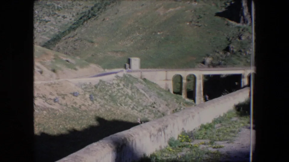 1971 Algeria 8mm frame showing a concrete bridge and road along a green hillside valley under bright daylight.