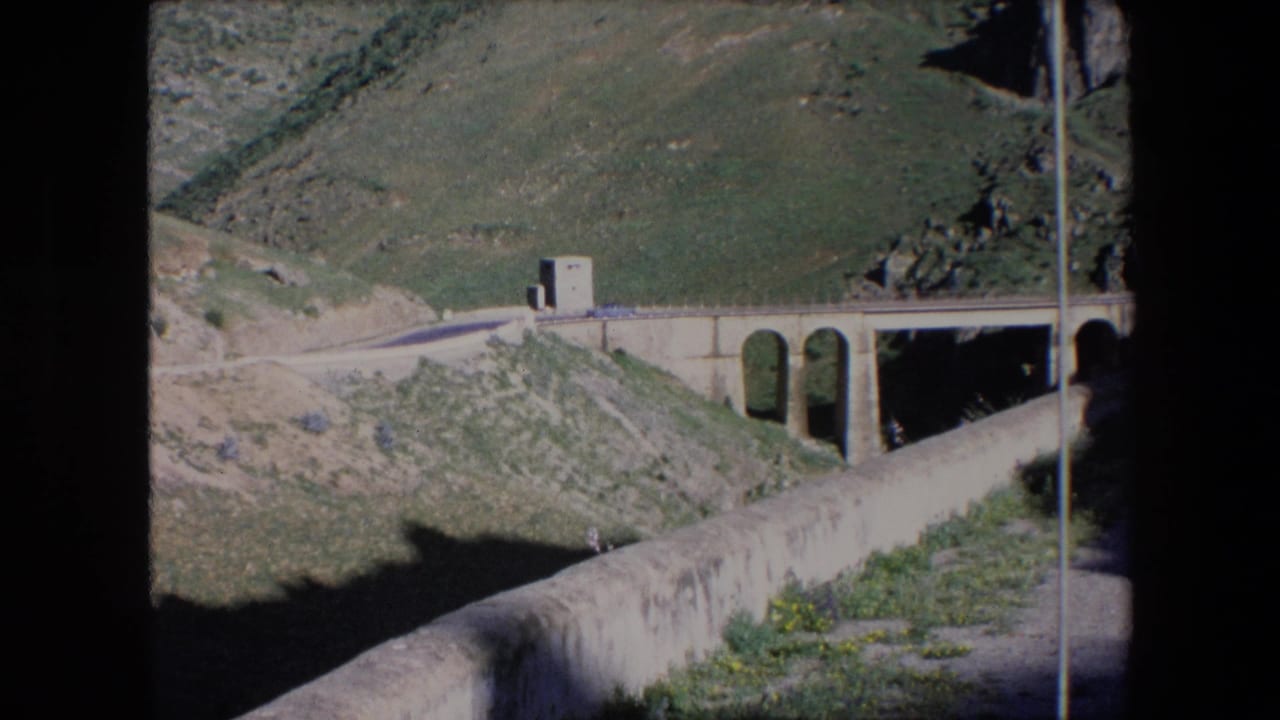 Small car on a mountain road near a stone-arch bridge and hillside.