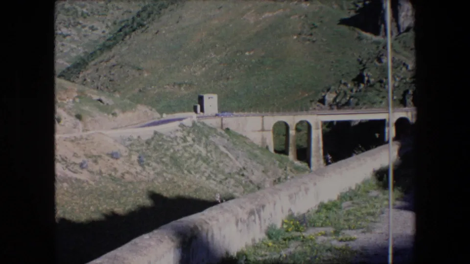 1971 Algeria home-movie frame showing a small car approaching or crossing a stone-arch bridge on a mountain road with green slopes behind it.