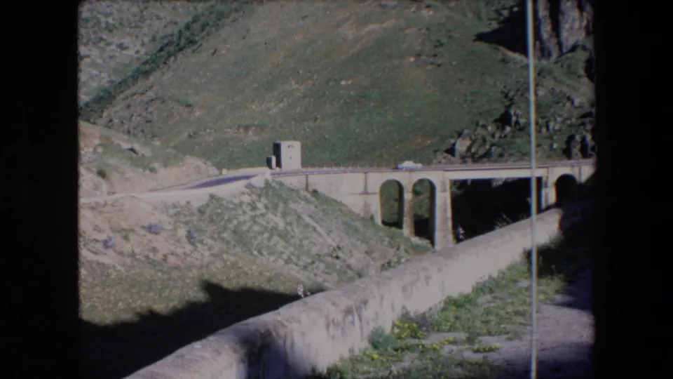 1971 Algeria home-movie frame with a small car on a bridge or roadside span, arched bridge openings, and rugged green hillside in bright light.