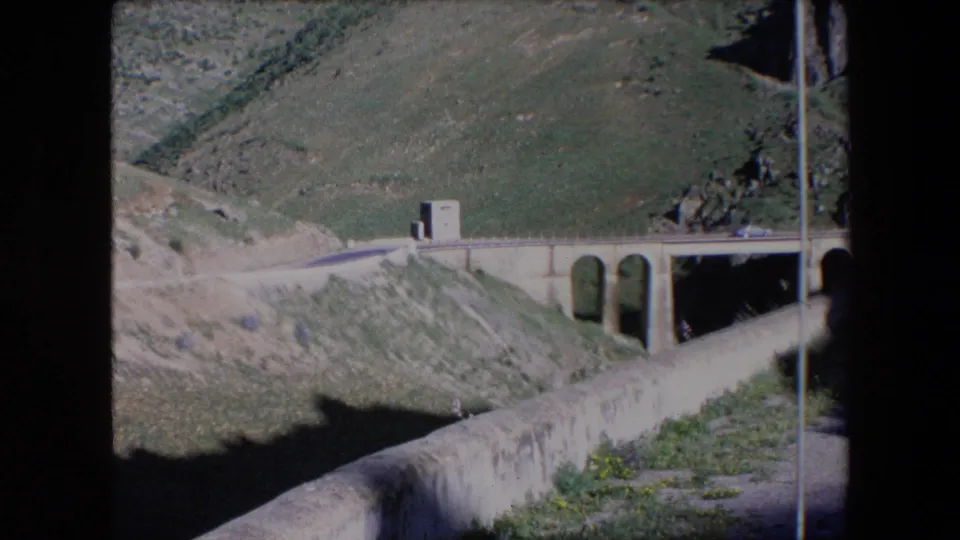 1971 Algeria home-movie frame showing the bridge and narrow road from a slightly shifted angle, with a small car, stone arches, and steep hillside terrain.