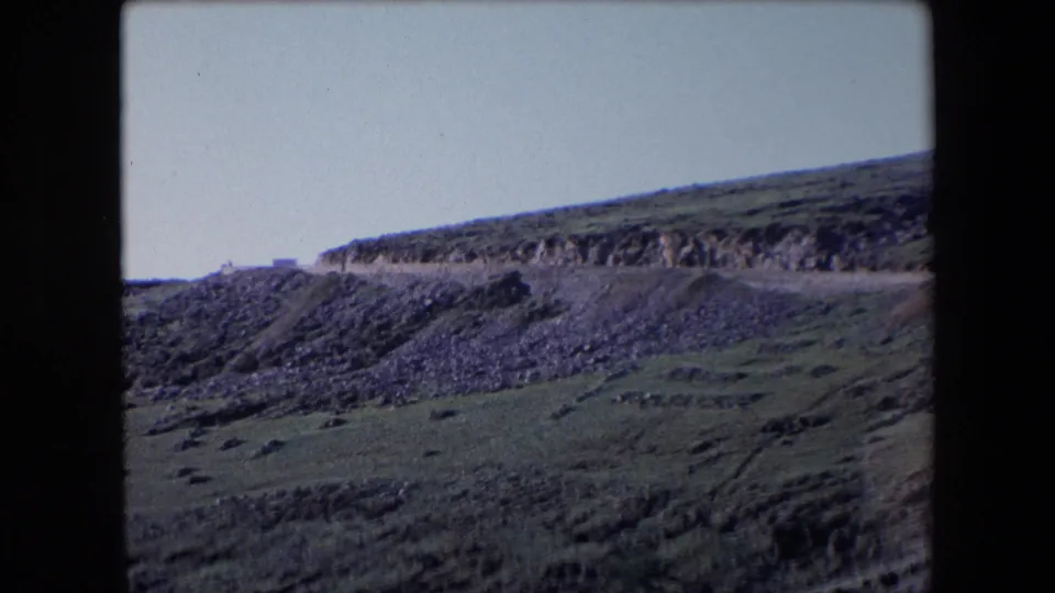1971 Algeria home-movie frame showing a distant car along a high mountain road, with rocky terrain, terraced-looking green slopes, and pale sky.
