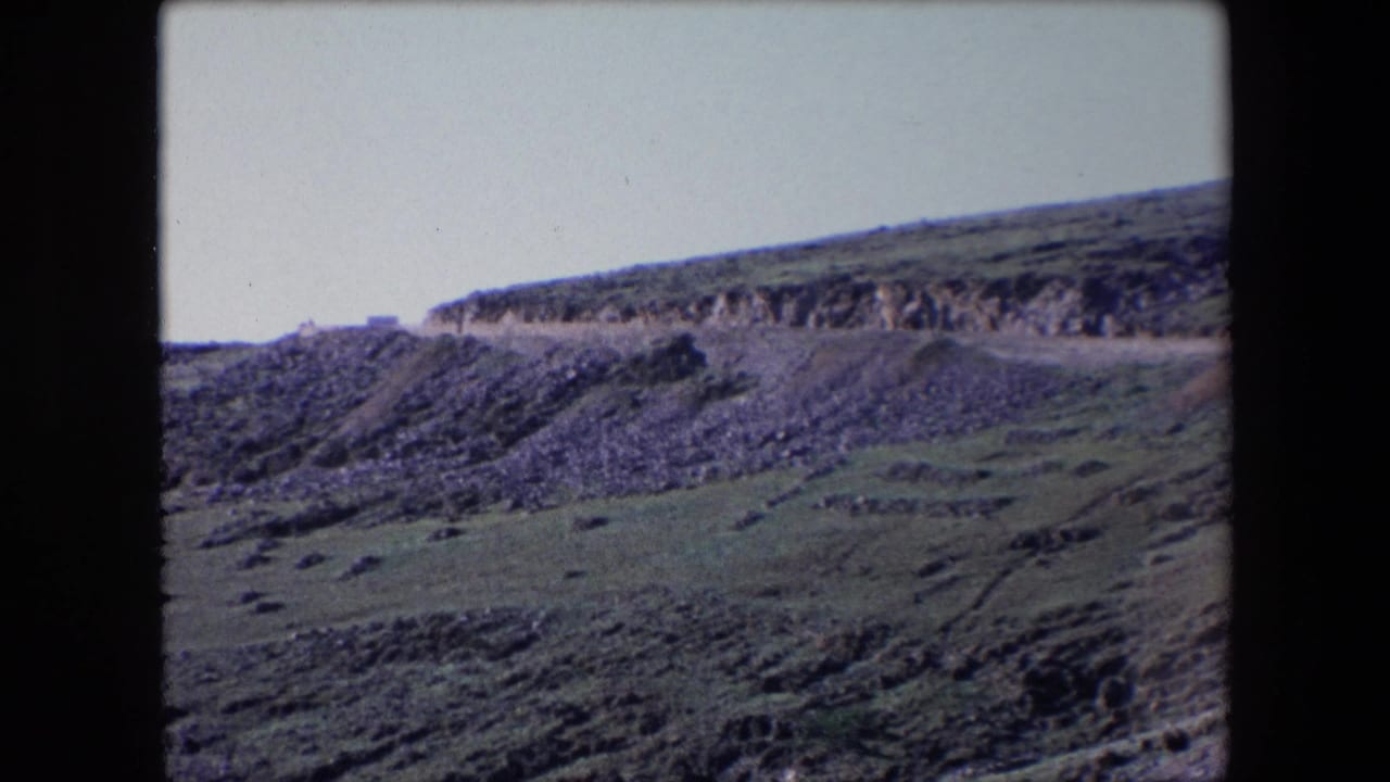 1971 Algeria 8mm frame showing a road cut across a steep rocky hillside with green scrub and a pale sky.