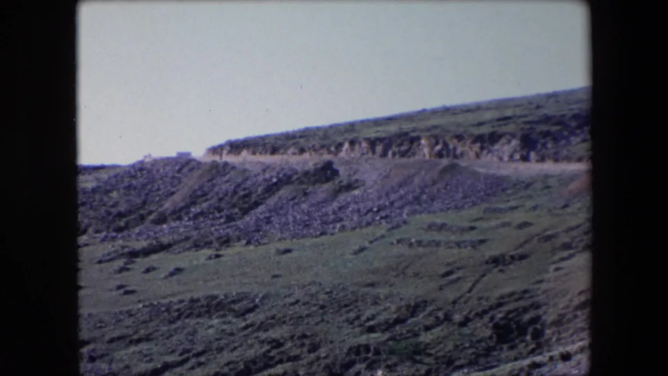 1971 Algeria 8mm frame showing a road cut across a steep rocky hillside with green scrub and a pale sky.
