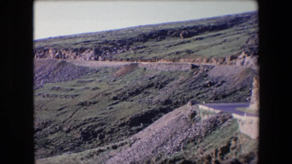 1971 Algeria 8mm frame showing a hairpin section of mountain road with a low barrier, scree, and scrub-covered slopes.