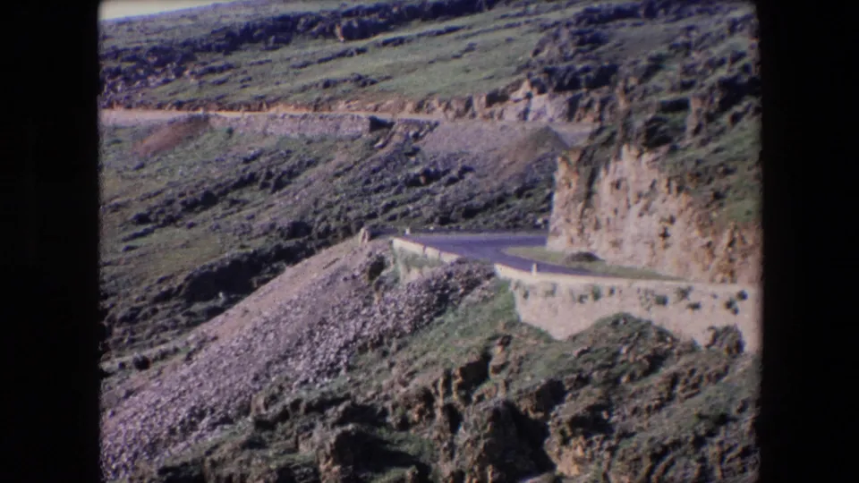 1971 Algeria travel frame showing the road bending around a rock face with a concrete wall and rugged hillside above.