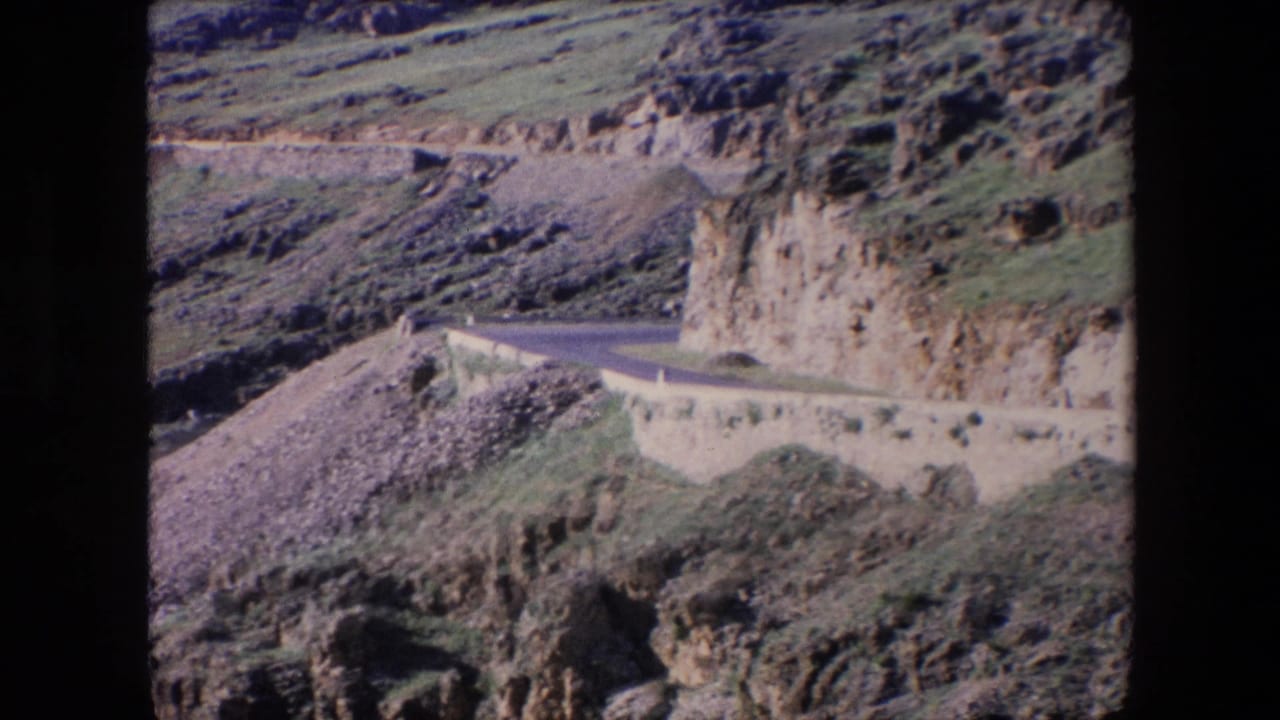 1971 Algeria 8mm frame showing a narrow paved road curving along a rocky hillside with a pale retaining wall and sparse green vegetation.