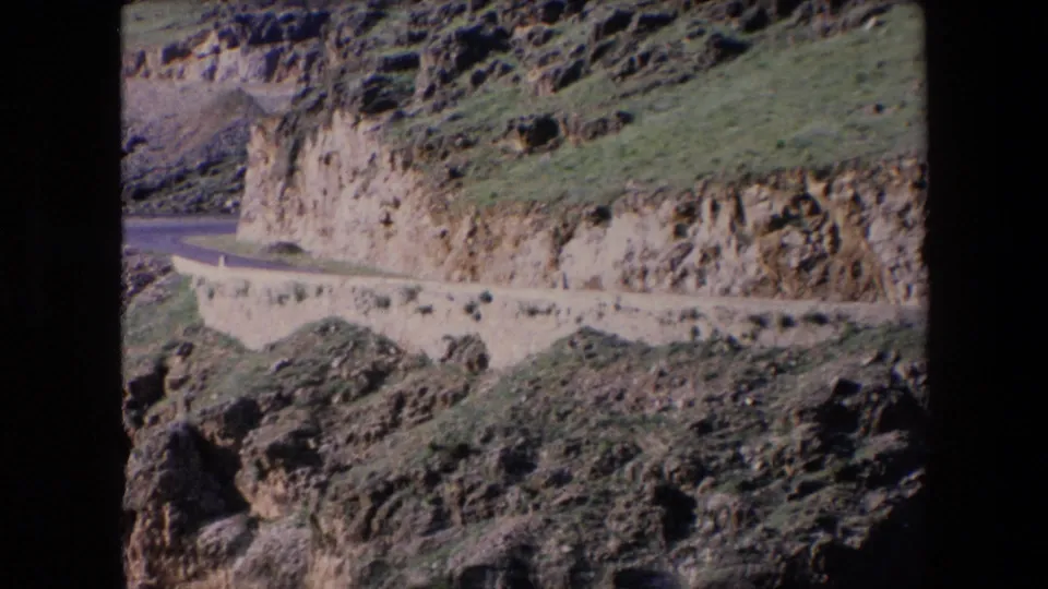 1971 Algeria 8mm frame with the mountain road stretching beside a cut cliff face and scrubby green slopes under bright light.