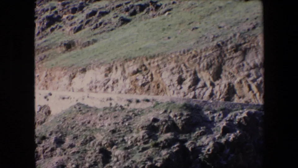 1971 Algeria 8mm frame showing the road partly lower in frame, bordered by a pale wall below a rugged slope and dark rocks in the foreground.