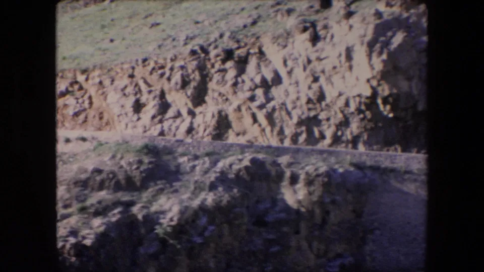 1971 Algeria 8mm frame concentrating on the eroded rocky cliff face and hillside above the road corridor, with the road edge partly visible below.