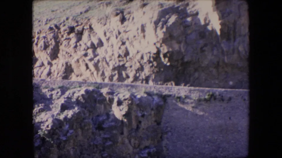 1971 Algeria 8mm frame showing a sunlit rocky hillside, exposed earth, shadowed stone faces, and a narrow dirt track across the terrain.