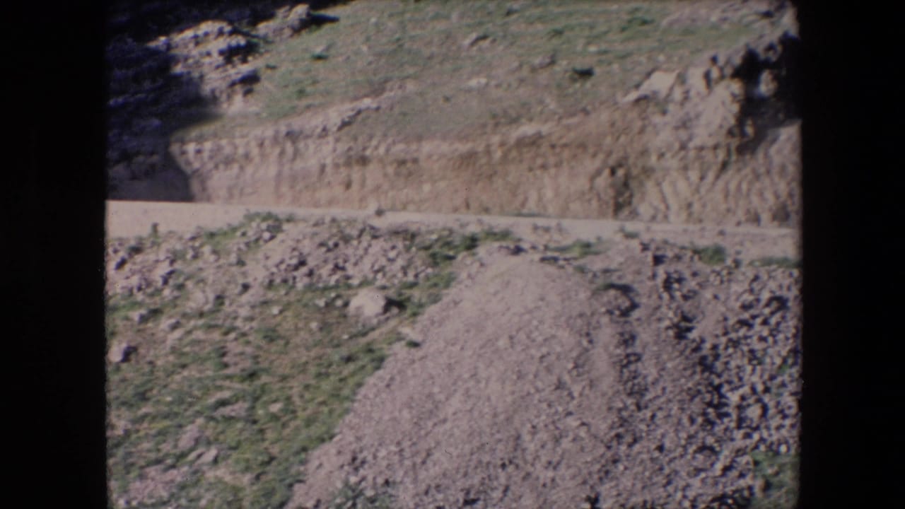 1971 Algeria 8mm frame showing a dry cut hillside, loose stones, a pale dirt road, and small green patches on rugged terrain.