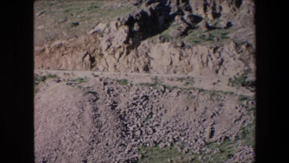 1971 Algeria 8mm frame showing pale exposed earth, scattered boulder piles, sparse vegetation, and a road or track crossing the rugged slope.