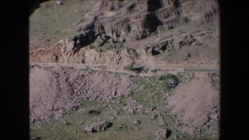 1971 Algeria 8mm frame showing a wider view of rugged eroded terrain, stone piles, pale sandy ground, and limited green vegetation under bright light.