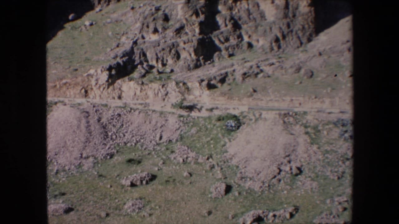 1971 Algeria 8mm frame showing a rocky arid slope, pale dirt path, scattered stones, and small patches of green vegetation.
