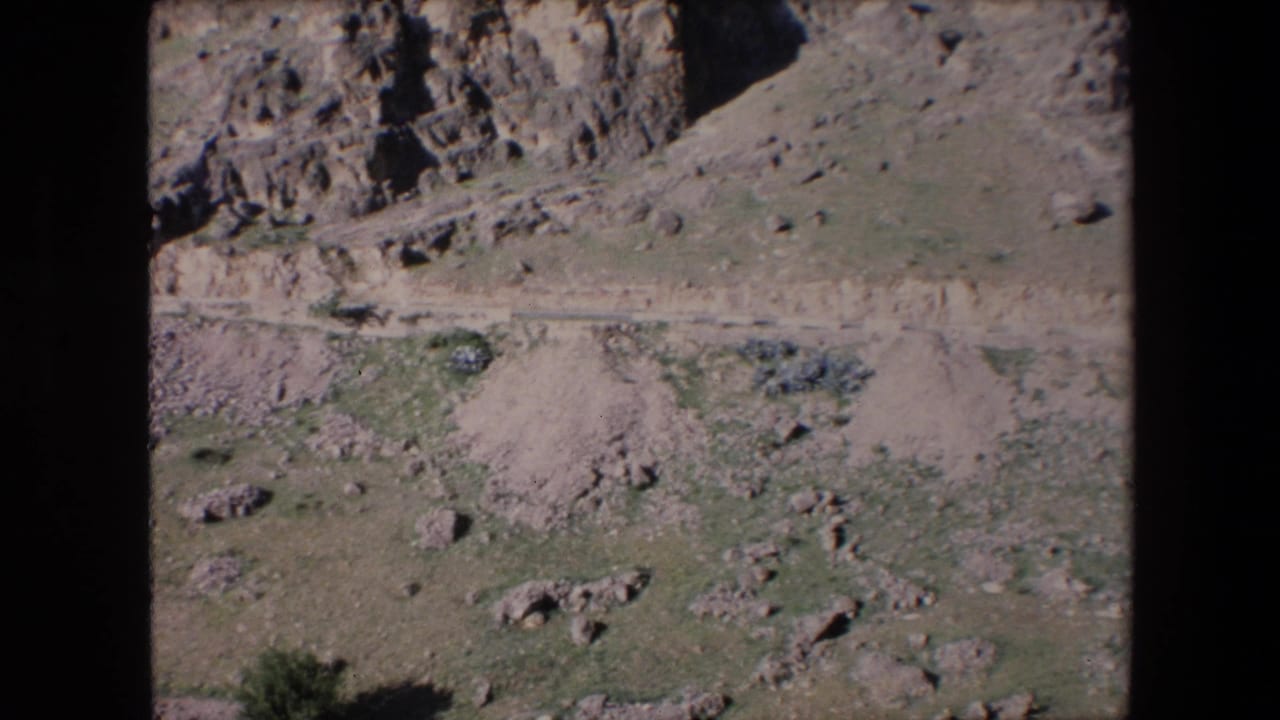 1971 Algeria 8mm frame with a barren rocky hillside, a narrow track crossing the scene, scattered boulders, and sparse scrub.