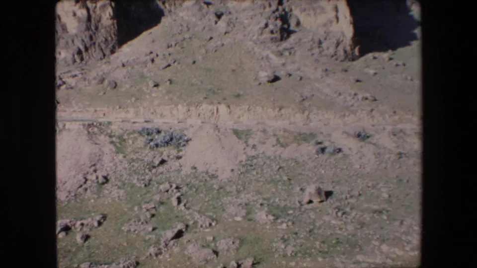 1971 Algeria 8mm frame showing rocky cliff edges, a pale dirt track, disturbed earth, scattered rocks, and sparse vegetation in an open dry landscape.