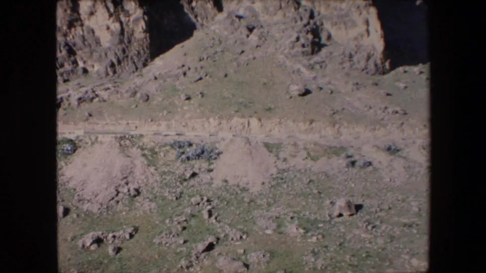 1971 Algeria 8mm frame looking across a dry rocky valley floor, with a narrow dirt path, scattered boulders, sparse green patches, and rough slopes behind.