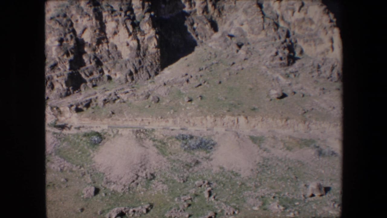 1971 Algeria home-movie frame showing a rocky arid hillside, scattered vegetation, and pale tracks or paths across the lower slope.