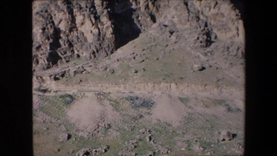 1971 Algeria home-movie frame showing a rocky arid hillside, scattered vegetation, and pale tracks or paths across the lower slope.