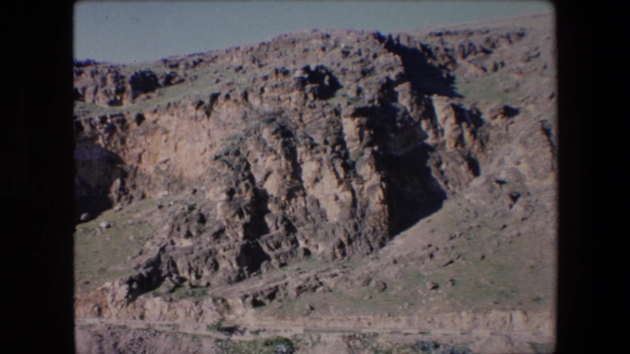 1971 Algeria home-movie frame with steep layered rock formations, a narrow path across the hillside, and open sky above the ridge.