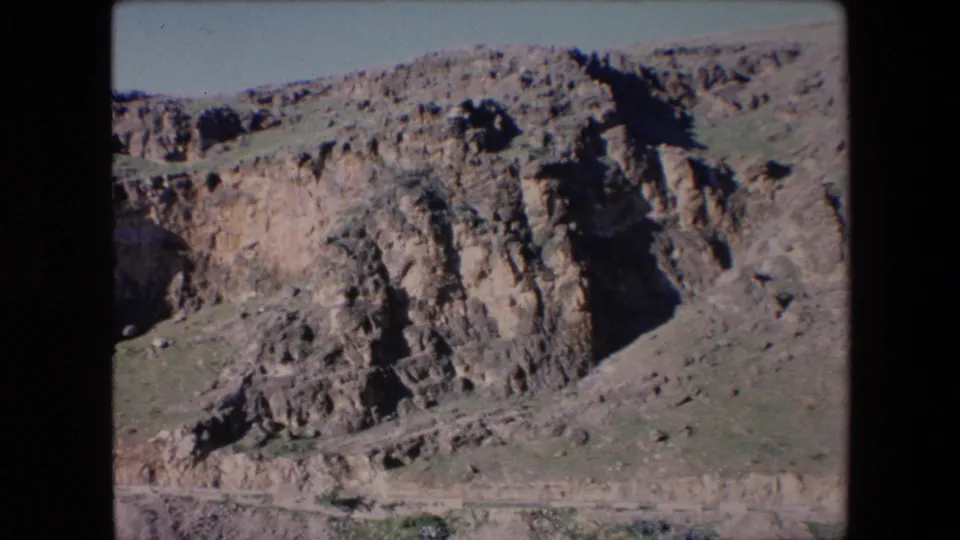 1971 Algeria home-movie frame with steep layered rock formations, a narrow path across the hillside, and open sky above the ridge.