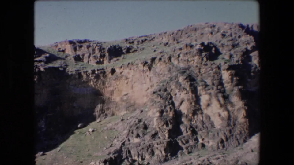 1971 Algeria home-movie frame looking across a rugged cliffside with uneven rock faces, sparse greenery, and a pale sky above.