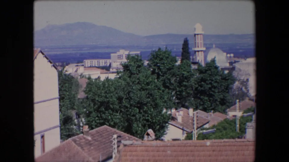1971 Algeria home-movie frame showing terracotta rooftops, trees, a white-domed mosque with a minaret, and mountains in the distance.