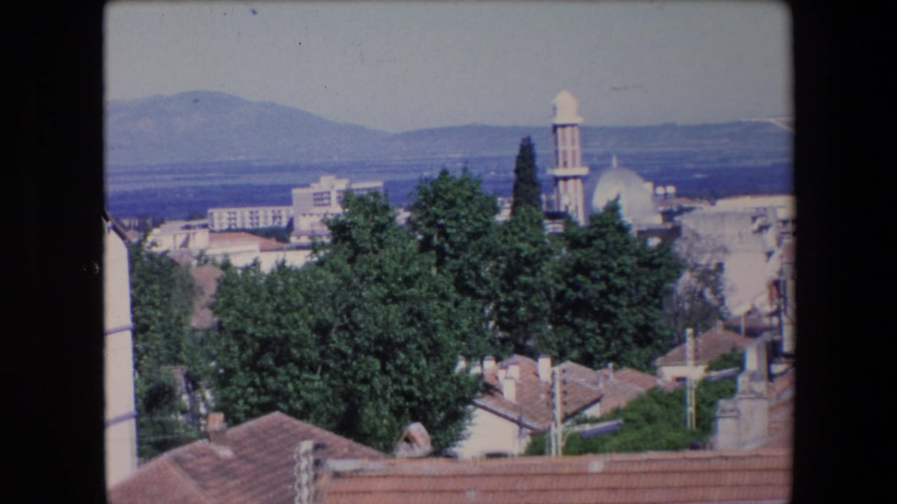 1971 Algeria home-movie frame looking over rooftops and trees toward a white-domed mosque with a minaret and hazy mountains beyond.