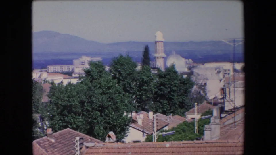 Soft 8mm frame from Algeria in 1971 showing an elevated town view with trees, tiled roofs, a mosque dome and minaret, utility poles, and mountains in haze.