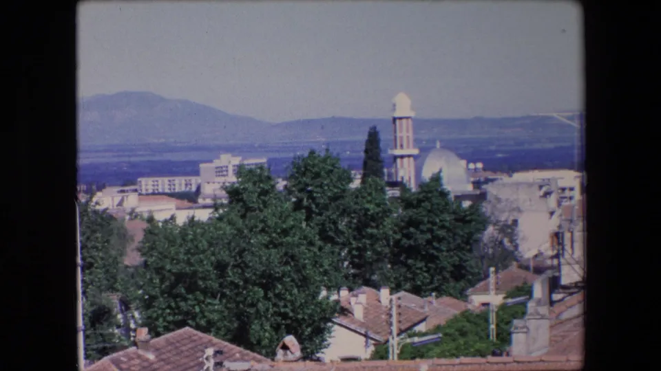 1971 Algeria 8mm frame holding on a townscape of red rooftops, green trees, a mosque minaret and dome, and a broad mountain horizon.