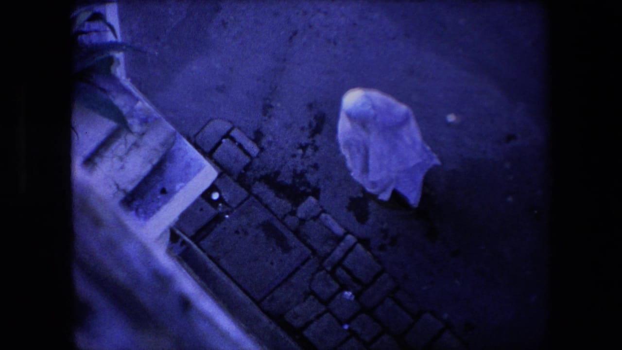 In 1971 Algeria home-movie footage, a person covered by a white sheet walks along dark pavement near a cobblestone edge and the side of a building.