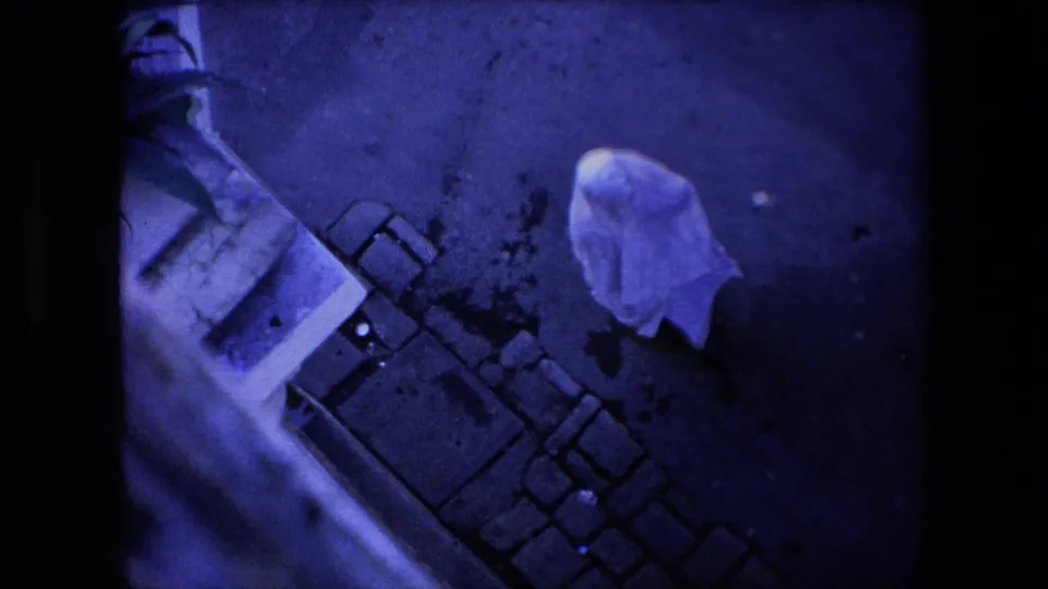 In 1971 Algeria home-movie footage, a person covered by a white sheet walks along dark pavement near a cobblestone edge and the side of a building.