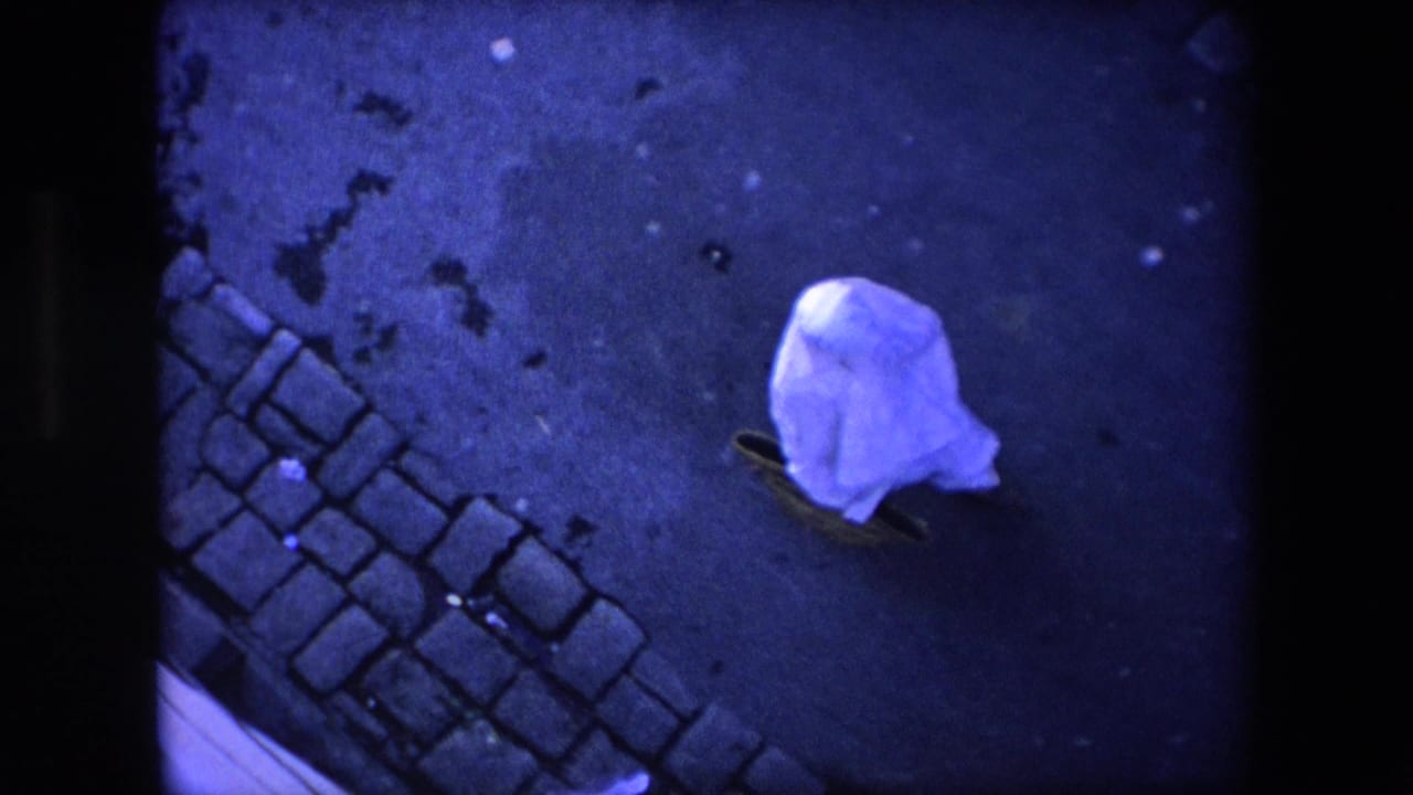 The sheet-covered pedestrian continues across a dark street surface near a strip of cobblestones in the Algeria 1971 home-movie clip.
