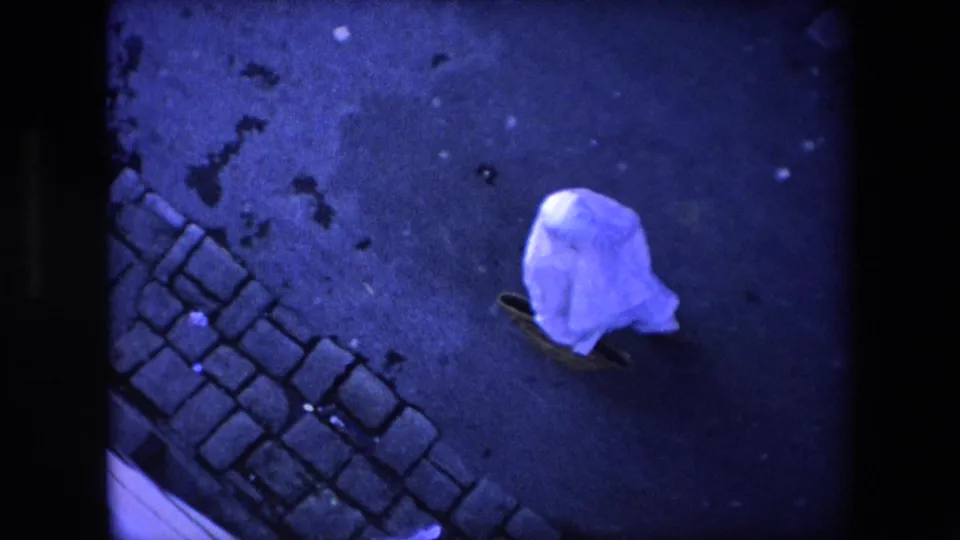 The sheet-covered pedestrian continues across a dark street surface near a strip of cobblestones in the Algeria 1971 home-movie clip.