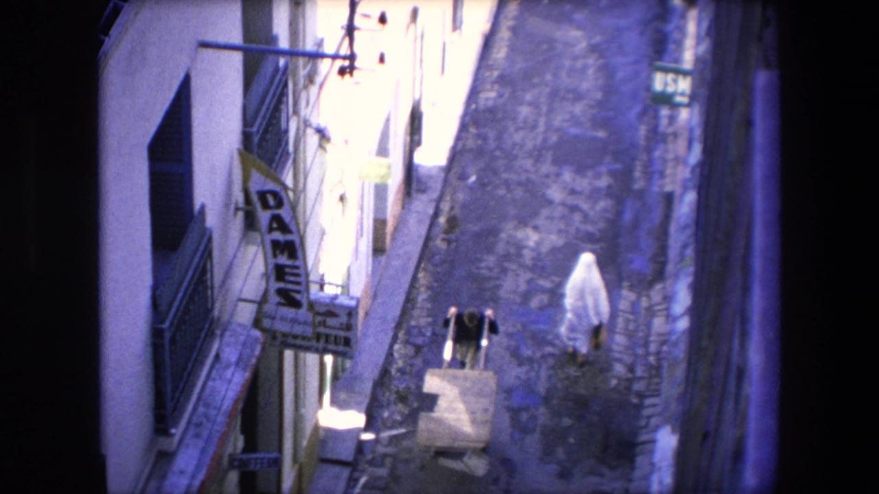 1971 Algeria home-movie frame showing a narrow cobblestone street from above, with a white-veiled woman walking away and another person pushing a cart near a vertical DAMES sign.