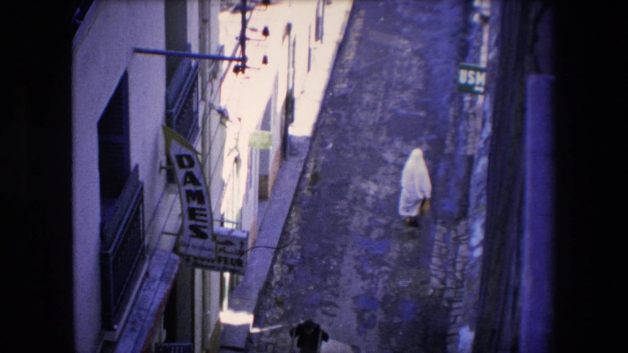 1971 Algeria frame from a high angle showing a white-veiled woman walking along a sunlit narrow street, with a partial view of another person and visible shop signage.