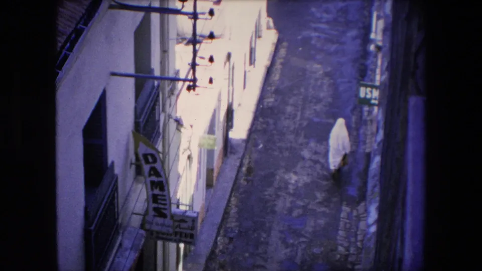 1971 Algeria home-movie frame showing the white-veiled pedestrian continuing along the narrow cobbled street, with balconies, a DAMES sign, and bright sun washing the upper street.
