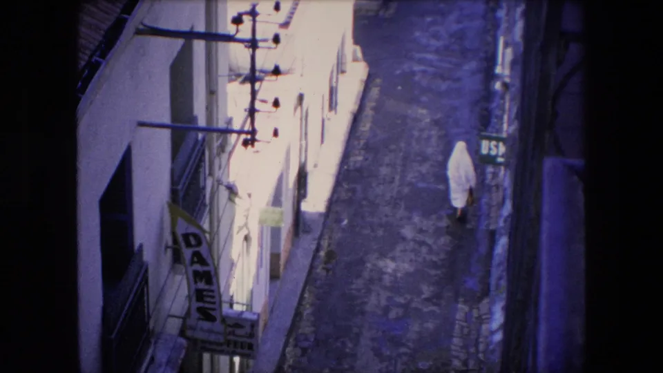 1971 Algeria frame showing a high-angle view down a narrow street as a white-veiled woman walks near the right wall, with visible DAMES and USM signage on adjacent buildings.