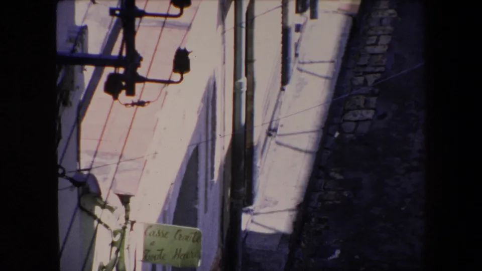 High-angle 8mm frame from Algeria in 1971 showing whitewashed walls, utility hardware, diagonal wires, a narrow cobblestone passage, and a partially visible sign near the lower edge.
