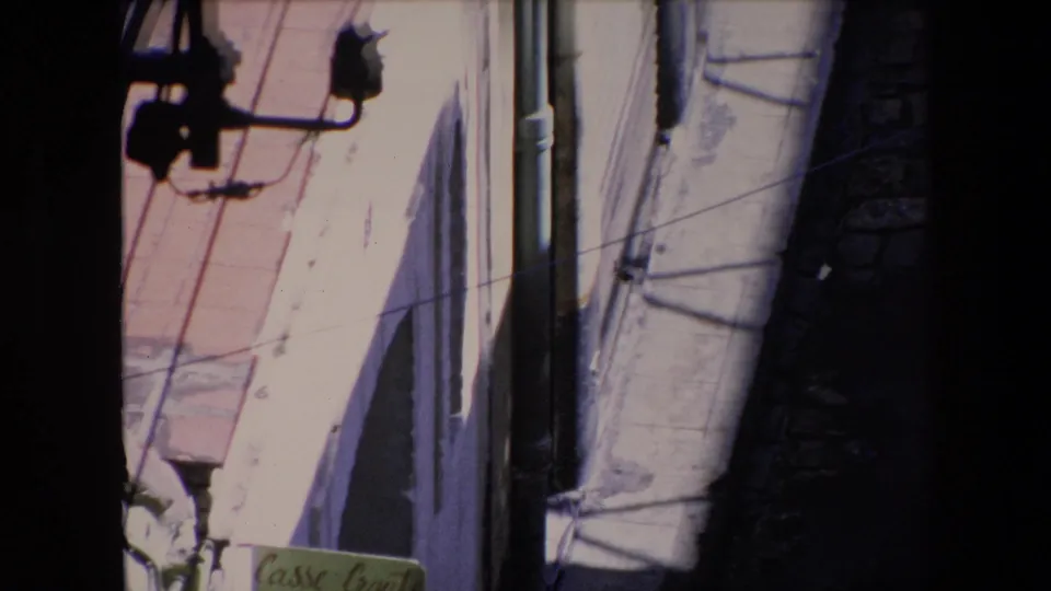 1971 Algeria 8mm frame showing a close high-angle view of a pale building facade, narrow alley edge, overhead wires, mounted fixtures, and deep shadow at the frame border.