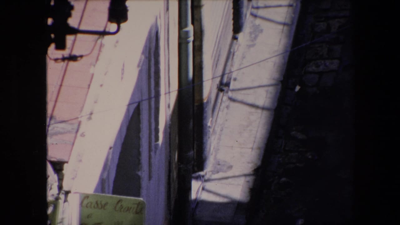 1971 Algeria home-movie frame looking down from above at white and pink stucco facades, vertical pipes, overhead wires, deep alley shadow, and a small green Casse Croûte sign near the lower edge.