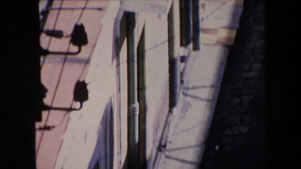 1971 Algeria frame from an elevated viewpoint showing pale stucco building surfaces, pink roof or balcony edging, overhead utility wires, a vertical pipe, and a narrow alley in heavy shadow.