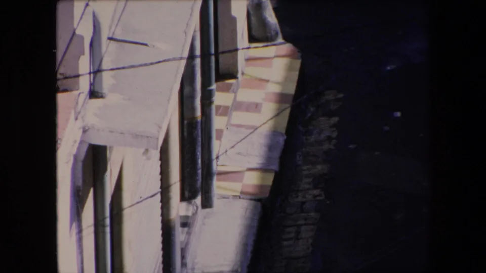 1971 Algeria home-movie still showing a tighter elevated angle on white stucco walls, dark vertical pipes, crossing wires, pink architectural trim, and the shadowed alley below.