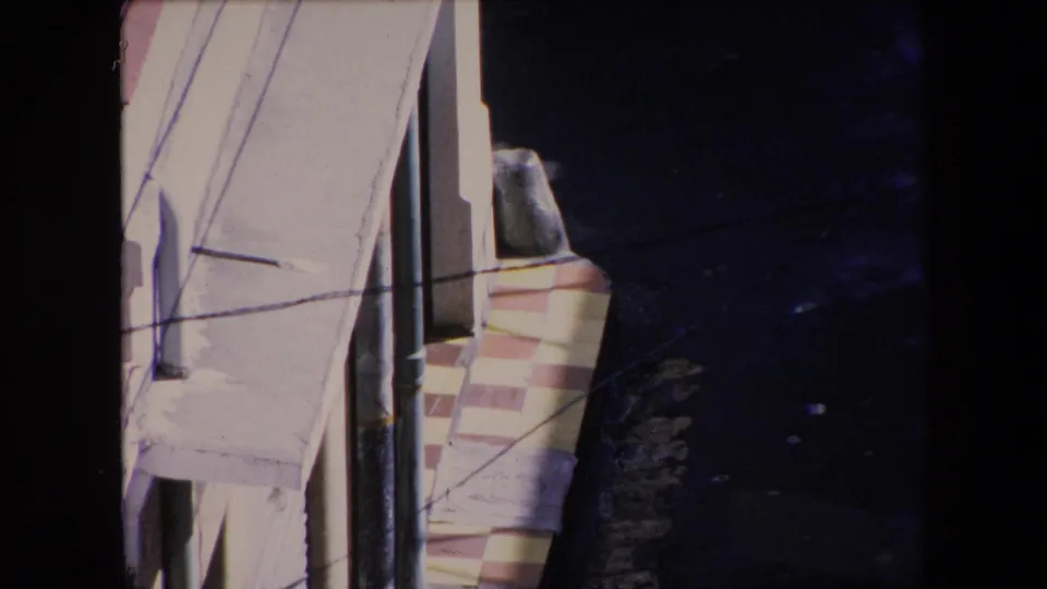 1971 Algeria frame looking downward along pale stucco facades with visible pipes, overhead wires, a window edge, pink trim, and a narrow alley receding into shadow.