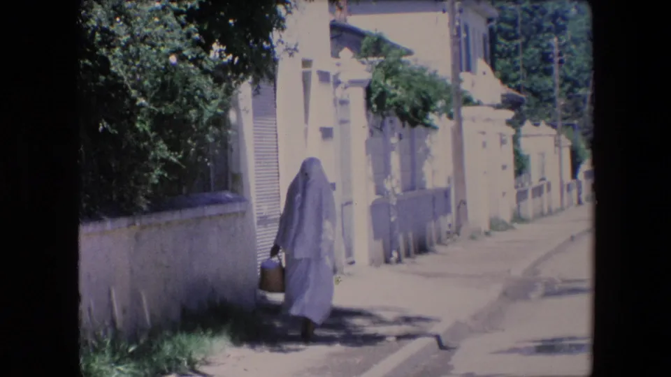 In metadata-supported 1971 Algeria, a woman in a white full-body covering walks along a quiet residential street beside a low wall while carrying a basket.