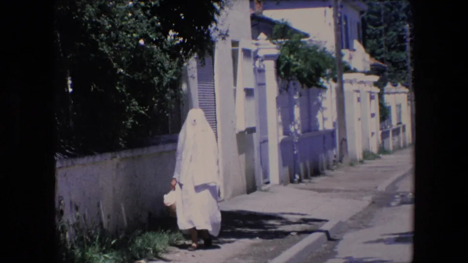 A woman in a white full-body covering approaches the camera on a sunlit Algerian residential street, with stucco buildings and trees behind her.